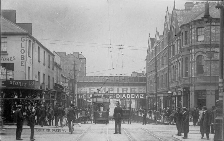 A black and white photograph of Bute Road, Cardiff. The photo is taken at a crossroads; a tram is heading towards the camera, as a carriage heads in the other direction.