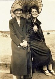 Black and white photo of Mable (left) and Doris (right), sisters on holiday in Bridglington. Here they can be seen resting on a wooden post on the beach, wearing long coats and heels.
