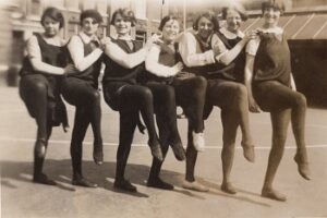Black and white photo of 7 netball players, posing with each right leg in the air.