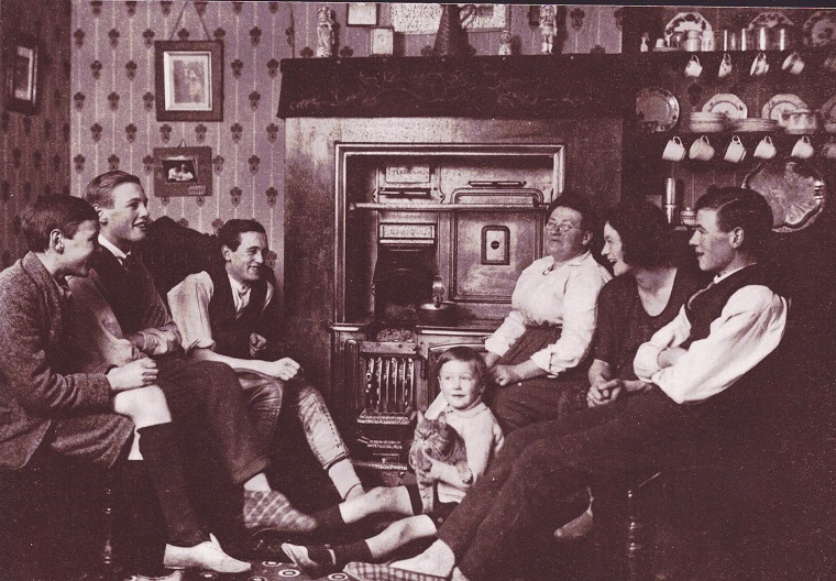 A black and white photo; 7 people - 6 children and their mother - sit around the hearth of their home. The youngest sits on the floor, holding a cat.