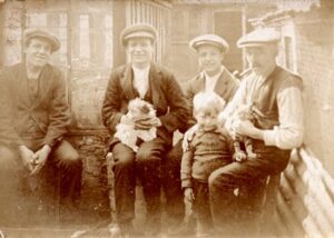 Sepia photo of three young men, a boy and their father, taken outside a house in North London. All except the young boy are sitting, wearing typical dress and flat caps. The middle of the three young men is holding a small terrier, while the father holds a cat.