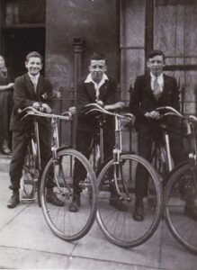 Black and white photo of three boys posing on bicycles on the pavement, mid-1920s.