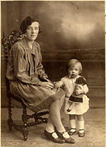A mother, sitting in a chair, with her young child standing next to her, holding a doll. c.1929.