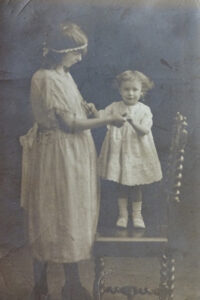 A toddler stands on a chair, facing the camera, as she holds hands with her aunt.