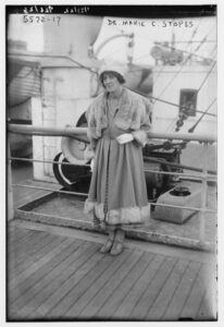 A black and white full-length photo of Marie Stopes, standing aboard a ship. Marie is wearing a coat with fur trim, a fur shawl, and hat.