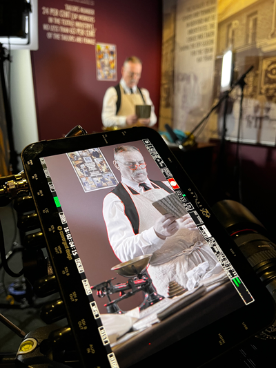 Behind the scenes shot filming the video series, depicting the shopkeeper character in the background and in the foreground, a digital camera screen showing the scene frame