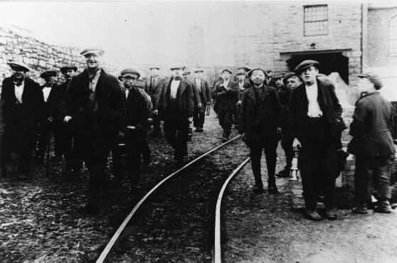 A photograph of Boldon Colliery Miners leaving post shift, with a track in the centre of the photo and all the men wearing flat caps and looking towards the camera