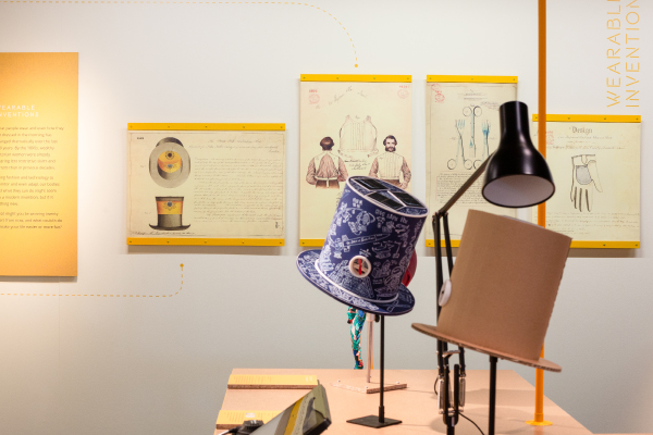 A photograph of the Spirit of Invention exhibition showcasing the Victorian designs on the wall and the modern interpretation of the ventilating top hat in the foreground