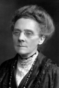 A black and white photograph of a woman dressed in Victorian clothes, wearing glasses, with a brooch at her neck and a necklace. 