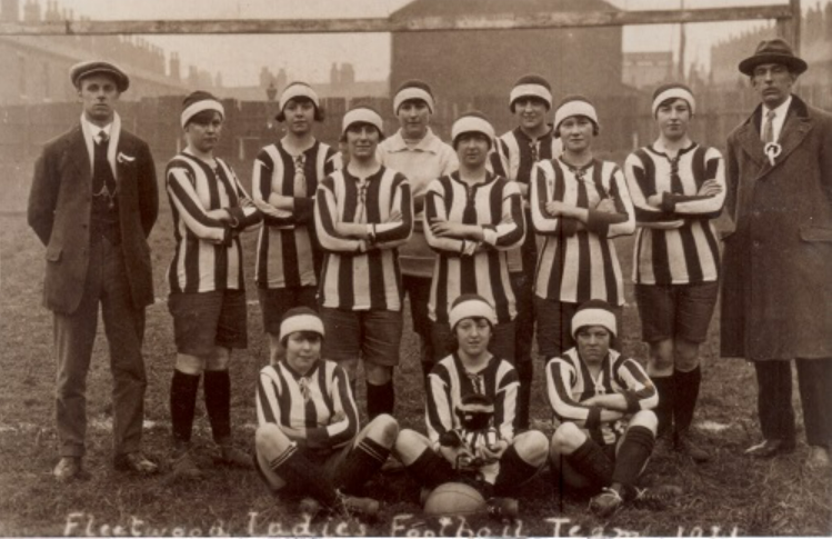 An image of a women's football team, dressed in uniform with 8 players standing, together with two men either end and 3 women sitting in front