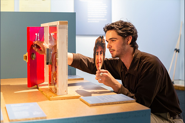 Photograph of model door pi in action in the exhibition. Someone holds the mask of Freddie's face up to a miniature door. 