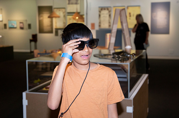 Photograph of child wearing XRAI glasses in the exhibition. The glasses are black and look like a pair of sunglasses.
