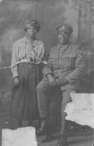 Young black couple pose for a photograph. The woman stands to the left of the photograph, while the man sits to the right and is dressed in military uniform