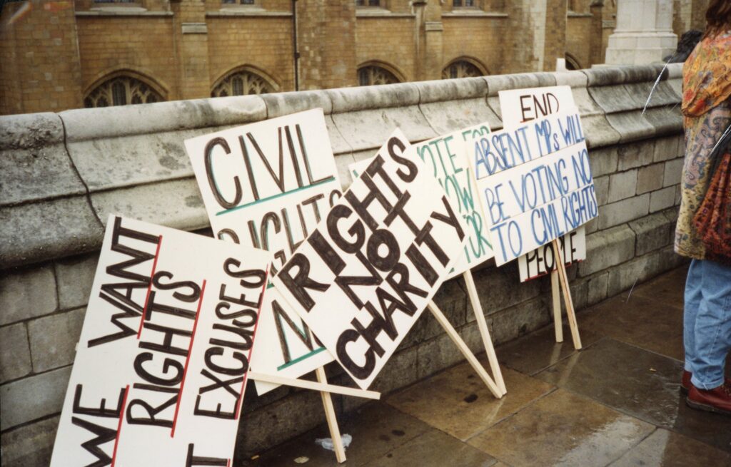 Various handwritten protest placards stood against a wall, with messages such as 'Rights Not Charity' and 'Absent MPs will be voting no to CML rights'
