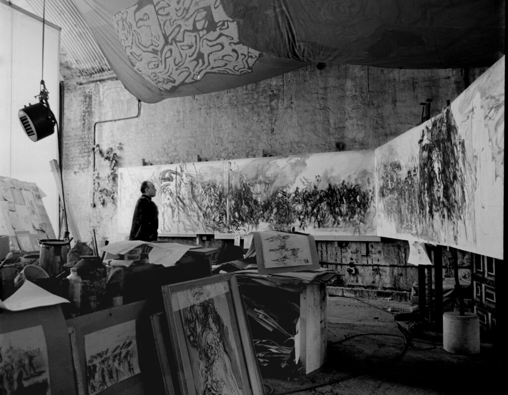Black and white photo of an artists studio, with various artworks visible, including a long landscape piece