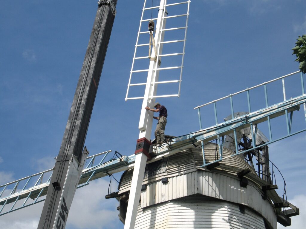 View of the top of a white windmill - a man in a flat cap is standing on the horizontal arm of the windmill, with a hand either side of the vertical arm, which is attached to a crane
