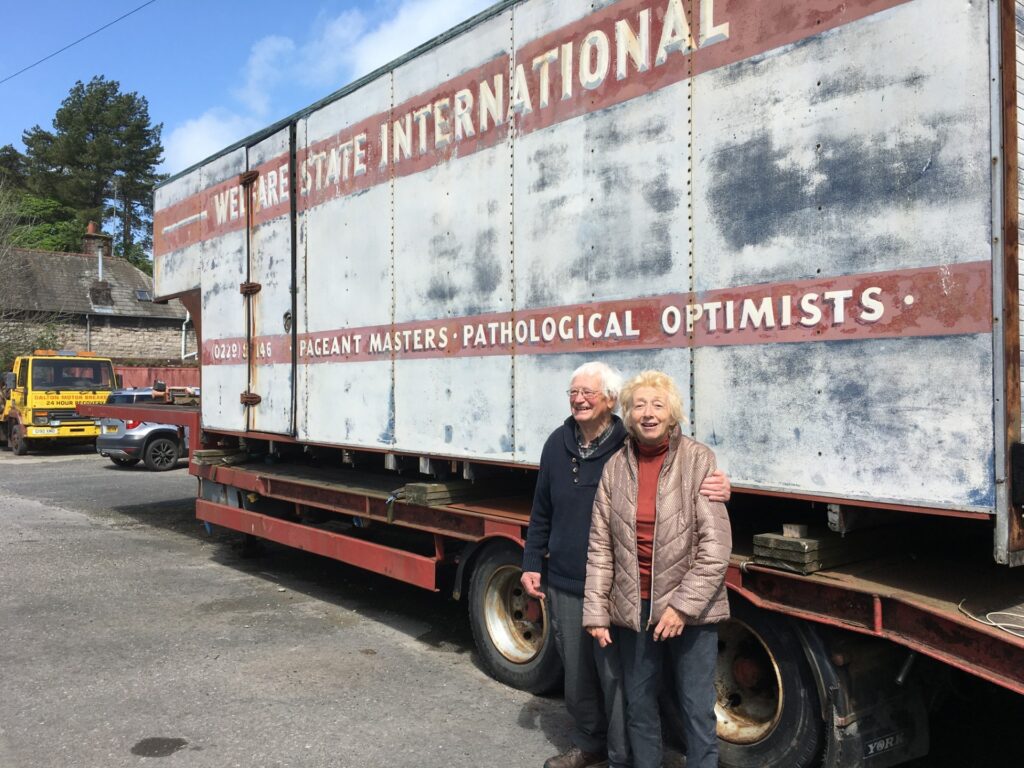 An elderly couple stood by a lorry trailer that has white-on-red signwriting that reads 'Welfare State International. Pageant Masters. Pathological Optimists.'