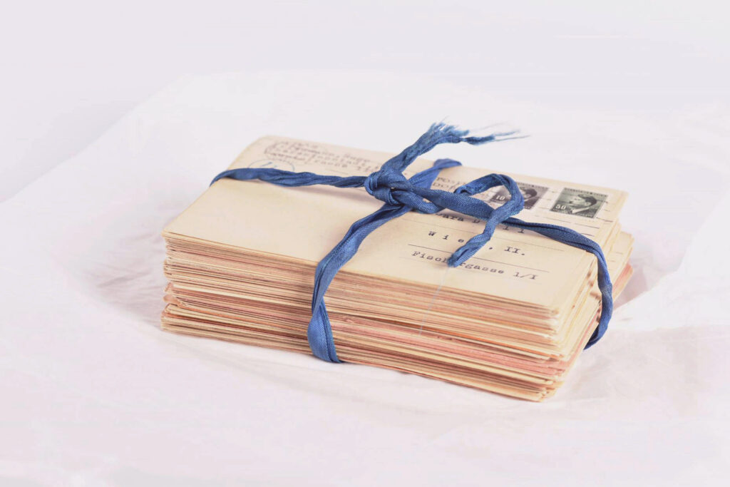 A stack of envelopes, bound with a navy blue ribbon. The address on the top envelope has been typed in German, and the two stamps on the top right of the envelope bear the profile portrait of Adolf Hitler.