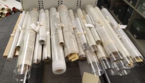 In a repository, dozens of large maps rolled up in plastic sheets rest on a table