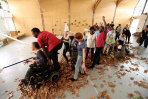 A large group of people parade through a line of brown fallen leaves in a large room with wooden walls.