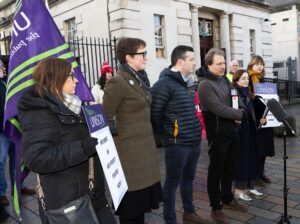 Several people are lined up outside a white building in front of a microphone stand. A man in the centre of the line appears to be speaking. Two women on either end of the line hold up 'Unison' signs.