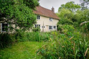 An exterior shot of a cream coloured house surrounded by trees, bushes and long grass.