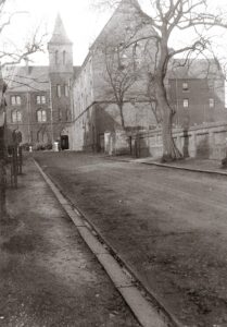 A black and white exterior shot of an old building with a leafless tree in the foreground.