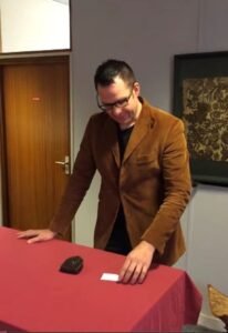 A man in a brown suede jacket stands over a table examining a small object.
