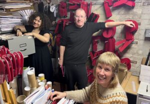 Three people in a small office are smiling for the camera, surrounded by filing cabinets and large red letter tiles.