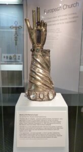 A golden model of a hand with regal sleeving, pointing upwards. This rests in a glass case with an information label entitled 'Shrine of St Patrick's hand'
