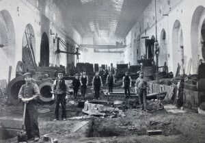 A black and white picture of around a dozen men working in an iron and steelworks factory. They all stare into the camera, some of them are holding work tools.