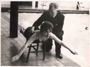 A black and white photograph of a woman balancing face down on a stool stretching her arms out while a suited man crouches next to her, adjusting her posture. They are beside an indoor swimming pool.