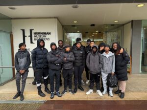 A group of a dozen boys and men, stand together outside a building with 'Horniman Museum & Gardens' engraved on the wall.