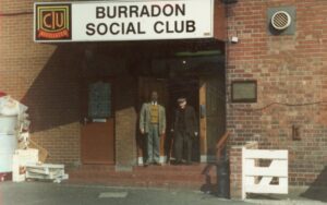 Two men in jackets and smart trousers stand at the entrance of a brick building under a sign that reads: 'Burradon Social Club'