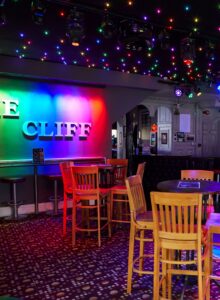 An interior shot of a bar illuminated with multi coloured ceiling lights. We can see two round tables with chairs, a sign on the wall that reads 'The Cliff', and a bar in the background.