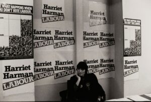 A black and white photograph of Harriet Harman sat at a desk. Behind her, there are many posters reading 'Harriet Harman Labour'