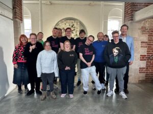 14 people stand together smiling for the camera in an open room with chipped brick walls. Several of the people wear 'This New Ground' branded t-shirts.