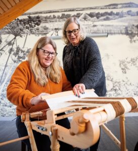 One woman supports another to operate a large wooden sowing machine