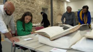 In a room with a cobbled brick wall, five people either sit at or stand over tables to examine records, ranging from large books to photographs and small cards.