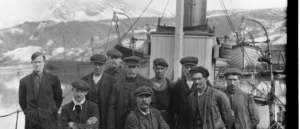 A black and white photograph of a dozen men stood on the deck of a ship against a backdrop of snowy mountains