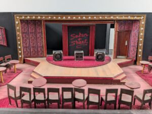 A set design model depicting a stage with three large sound speakers. Much of the décor (carpets, wallpaper etc.) is made of crimson and burgundy reds. 