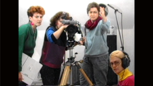 Four people are gathered around a film camera, tripod and boom mic. One appears to be directing the off-screen action, one operates the camera, one wears headphones and another appears to carry a clipboard. The image has a 1980s VHS style texture.