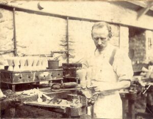 A faded black and white photograph of a man, wearing an apron, stood in a tool workshop making brushes.