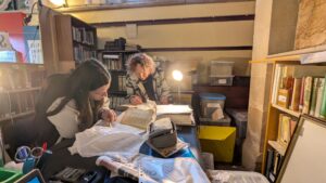 Two researchers study paper documents, under lamp light, in an archive space with lots of boxes and book shelves.