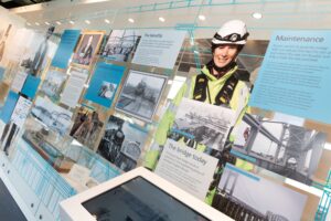 An exhibition display featuring information boards and photographs relating to Tamar Crossings (most prominently a woman wearing a high-vis jacket and a safety helmet).