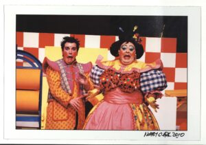 Two performers wearing colourful costumes stand in front of red and white chequered scenery pulling colourful expressions. One of the performers appears to be styled like a pantomime dame.