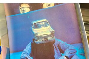 A photograph of someone sitting down wearing blue overalls and the model of a car on their head.