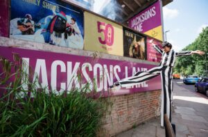 A performer on stilts walks past a banner reading 'Jackson Lane' and a series of posters for shows.