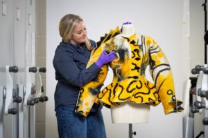 A curator examines a yellow and black jacket on a headless mannequin. 