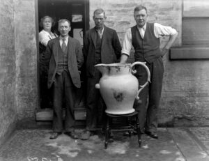 A black and white photograph of four men posing alongside an enormous jug.